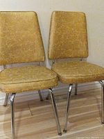 Two vintage kitchen chairs with harvest gold vinyl upholstery and chrome legs side by side against wall