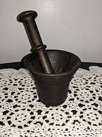 Photo showing cast iron mortar and pestle set placed on a crocheted doily, mortar upright with pestle inserted.