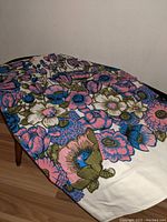 Close-up side view of the floral pattern on the vintage tablecloth draped over a table, showing large pink and blue flowers with olive green leaves and white background.