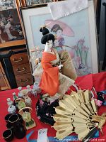 Porcelain Geisha doll in orange kimono in front of two framed needlepoint Asian art pieces, along with sake set cups and bottles, small ceramic figurines, and a large decorative golden fan shaped ornament.