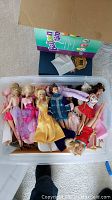 Top view of plastic bin open, showing various Barbie dolls dressed in colorful outfits including pink, purple, yellow. Bratz doll also visible. Box lid with Barbie branding partly visible.