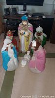 Overview of six blow mold Nativity figures arranged standing and lying on carpet near entertainment unit.
