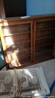 Wooden wall hanging storage cabinet with glass doors and visible shelves inside. Cabinet has scratches on surface.