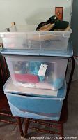 Three stacked plastic storage containers filled with assorted items such as linens and doll accessories.