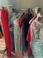 Photo shows metal clothes rack with various womans dresses hanging, some in plastic garment bags. An ironing board and red outdoor umbrella bag visible next to rack.