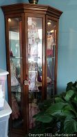 Front view of corner curio cabinet showing wood finish, glass door panes with interior display and dolls inside, brass pull handle visible.