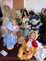 Photo showing a group of approximately 12 porcelain head dolls in varied outfits including blue dress and large hat, pink dress, and vintage lace-clad doll with bonnet.