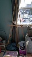 Photo showing wooden tripod easel standing by window with items piled at base.