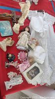 Wide shot of porcelain dolls, figurines, baptism outfit and bonnet on red surface.