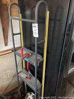 Full view of the hand truck showing gray metal frame with yellow edge guards and red handles, positioned near a black appliance.