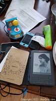 Photo of all devices on table: Kindle e-reader displaying text, owner's manual, blue iPod Nano, three cell phones, green Ms. Pac-Man joystick.