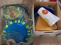 Photo showing plastic plates, a plate with a parrot design in plastic packaging, a blue bowl and spiral metal flatware holder, alongside a box containing a citrus design pitcher and yellow woven mats.