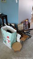 Photo of miscellaneous items grouped including small wooden step stool, white gift bag with contents, framed picture frames leaning against wall, walking sticks, and other framed art.