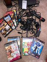 Full lot overview showing the PS2 slim console, multiple game cases including SingStar and American Idol, two wired microphones, two wired controllers, and tangled cables.