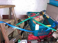 Two metal saw horses strapped together with blue tie, with plastic basket containing painting supplies on top, long-handled roller resting on top.