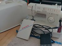 Singer sewing machine with white hard plastic case, foot pedal, and instruction manual on the floor.