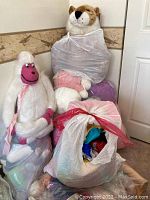 Photo showing large white and pink ape stuffed animal seated on top of plastic bags filled with doll clothes and other stuffed animals, a large tiger stuffed animal standing behind.