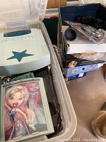Photo showing Bulldog media security lock and Bratz doll carrying cases in plastic storage bin, with multiple game and toy boxes stacked to the right.