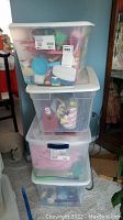 Four large clear plastic storage tubs stacked, filled with various toys and dress-up items.