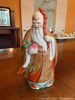 Front view of a hand painted ceramic figurine showing the smiling man in a gold robe holding a peach and red staff.