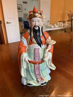 Front view of hand painted Chinese ceramic figurine showing the man with a black beard and orange robe, holding items in both hands.