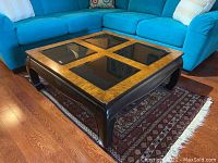 Oblique top-front angle of square coffee table with smoked glass panels and burled wood frame, placed on oriental rug in front of a turquoise sectional sofa.
