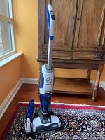 Full view of cordless Hoover Floormate Jet floor and rug cleaner upright on a patterned rug in front of wooden furniture and window.