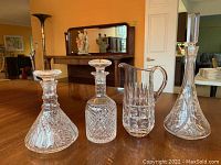 Three cut glass decanters and one glass water jug displayed on wooden table with furniture in background.