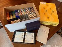 Box filled with multiple music CDs and boxed sets on a wooden table, showing variety of jewel cases and box collection covers.