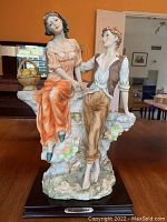 Front view of porcelain figurine of a young woman in an orange dress sitting on rock ledge and young man standing beside her, holding her hand.