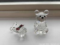 Both Swarovski crystal teddy bears on a white surface, one smaller with plaid ribbon, one larger sitting upright.