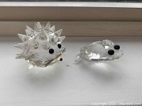 Two Swarovski crystal figurines placed side by side on a white surface under natural light, showing both the hedgehog and frog front views.
