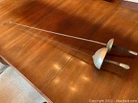 View of two fencing foils lying side by side on a wooden table, showing overall length and shape.