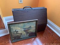 Framed litho print and vintage cleaning kit wooden case leaning against wall, visible wear on case and print frame