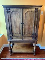 Front view of vintage wooden cabinet showing two paneled doors, two drawers below, and bottom open shelf on turned legs with carved accents.