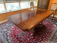 Dining table with removed leaf, showing polished wood surface and beveled edges, photographed indoors on a red patterned rug near windows.