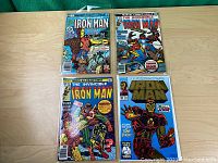Four Iron Man comic books laid flat on a light wooden surface with clear protective covers. The comics show various colorful covers with Iron Man and associated characters in action poses.