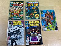 Five Iron Man comic books arranged on a light wood floor showing their colorful front covers and various titles from different issues.