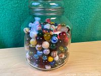 Full view of the glass jar filled with colorful vintage marbles on wooden surface and green background.