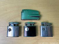 Four vintage Ronson lighters laid out on a wooden surface: three chrome finish with ribbed design and one green enamel lighter.