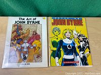 Two graphic novels side by side on wooden surface with green background. Left: The Art of John Byrne Volume One, Right: Focus on John Byrne by Fantagraphics Books.