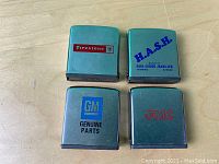 Four vintage metal Zippo tape measures with advertising logos from Firestone, H.A.S.H., GM Genuine Parts, and FMC on wooden surface.