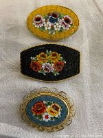 Three vintage glass mosaic pins laid flat on surface, showing floral designs with yellow, black, and blue backgrounds respectively.