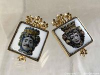 Pair of Italian gold leaf tile earrings with detailed faces and gold floral accents against a beige background.