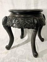 Front side view of carved dark wooden Asian foot stool showing detailed floral and leaf carvings with curved legs ending in curled feet.