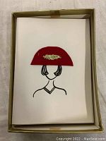 Top-down photo of a single blank card featuring a woman's head illustration wearing a small red felt hat with gold glitter decoration, showing detail and design of the card.