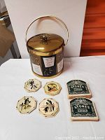 Front view of ice bucket with hinged lid and handle. Bucket decorated with cocktail recipes and embossed illustrations. Eight vintage cardboard coasters laid out in front.