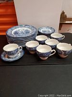 Photo showing eight 9-inch plates stacked and grouped teacups with saucers around them, all with blue scenic and floral Liberty Pattern design