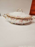 View of the entire covered porcelain serving bowl with pink floral pattern and gold trim, shows curved handles and scalloped lid edges.