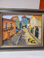 Framed oil painting showing the street scene with colorful buildings, vintage car, and signature on lower right.